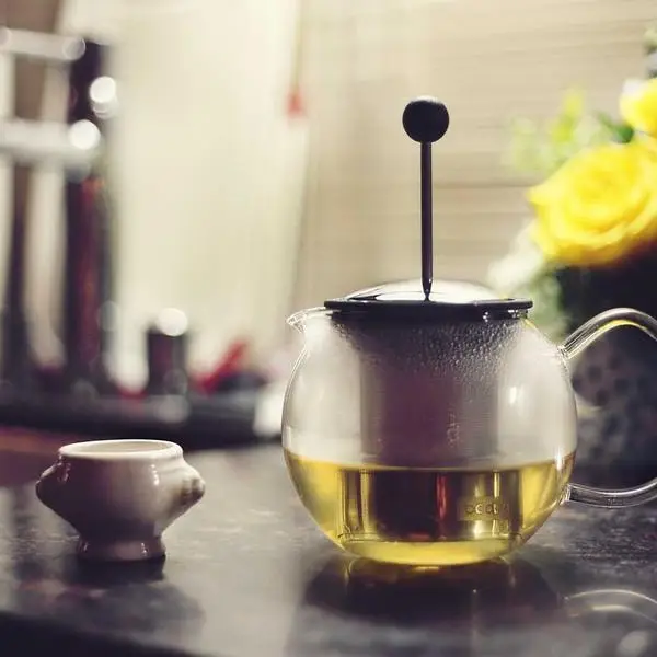 Glass teapot releasing steam as tea steeps on a kitchen counter, small cup and yellow flowers blurred in background.